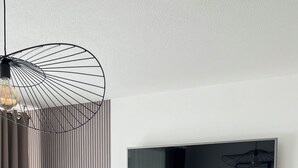 Un salon moderne avec un téléviseur fixé au mur, un miroir rond, un bureau avec une chaise et un luminaire suspendu original.