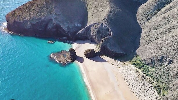 An aerial view of a coastal area with a beach, rocky cliffs, and clear blue waters.