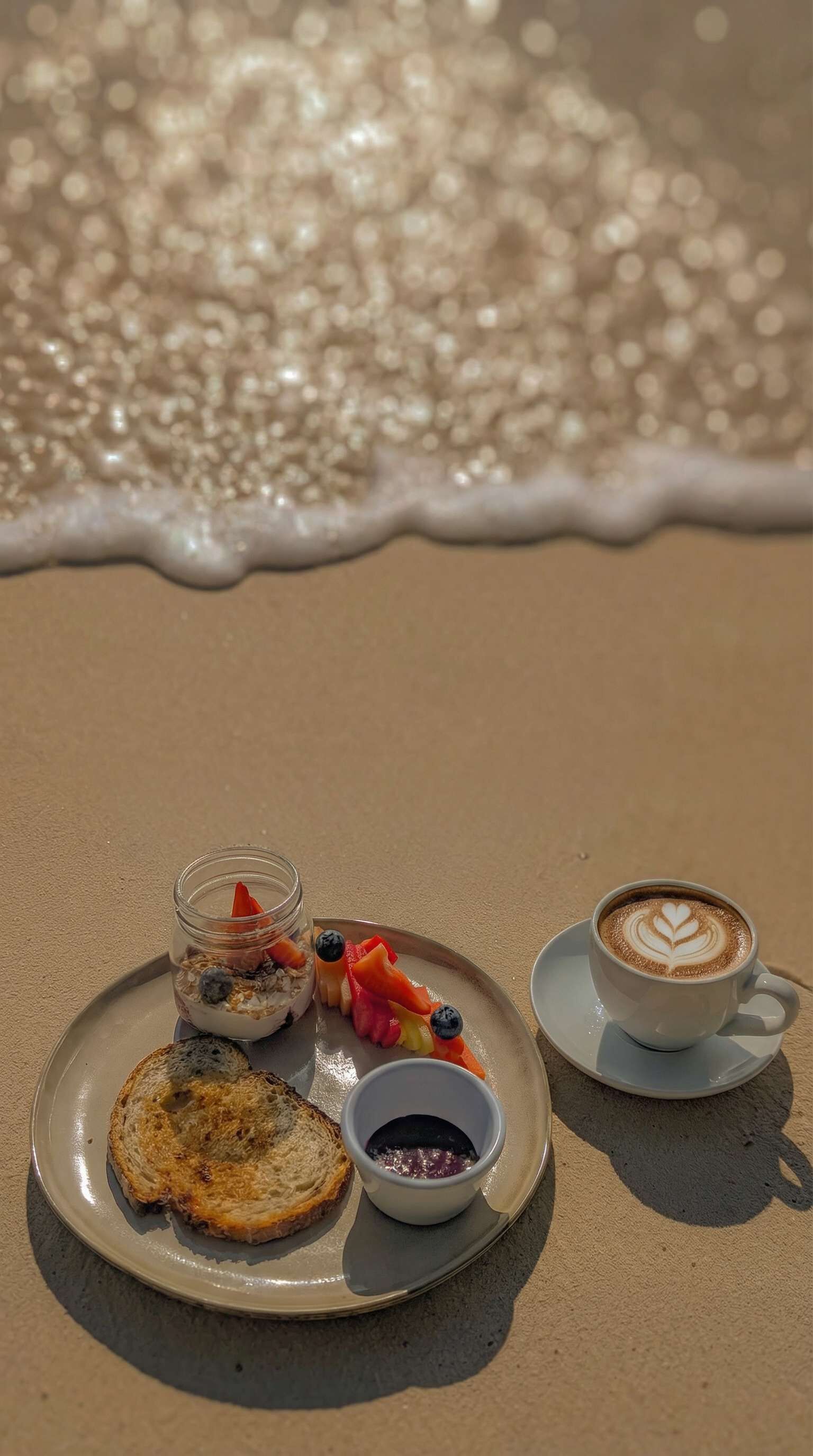 Un petit-déjeuner servi sur la plage, composé de toasts, de fruits, d’une tasse de café et d’un petit pot de confiture.