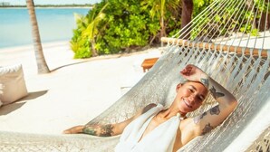 A person relaxing in a hammock on a beach with a thatched roof, palm trees, and a clear view of the ocean. - La Casa: 3 Bedroom Suite (Tulum)