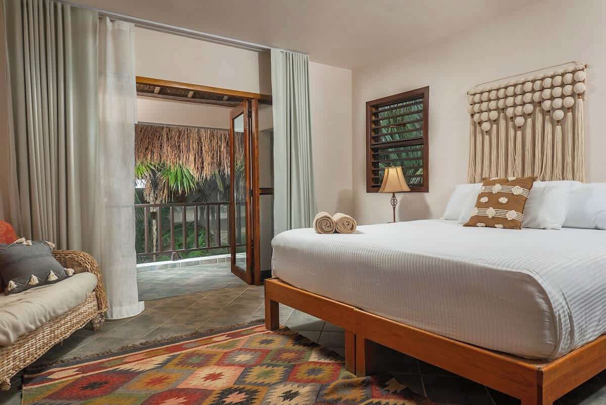 A hotel room with a bed, a sofa, a patterned rug, a window with curtains, and a view of greenery outside.