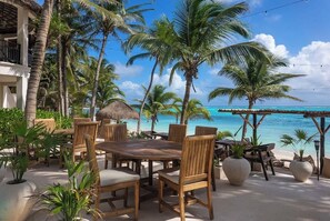 An outdoor dining area with wooden tables and chairs, surrounded by palm trees and a view of the ocean. - La Casa: 3 Bedroom Suite (Tulum)