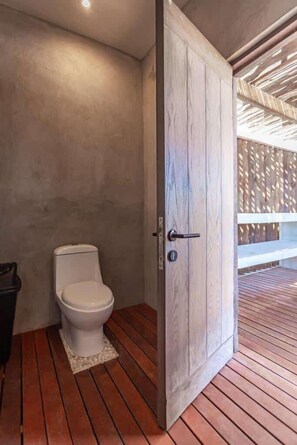 A modern bathroom with a wooden floor, a white toilet, and a large wooden door. - Tankah Bay Beachfront 3BR with private Rooftop Sea Views | Sandy Beach (Tulum)