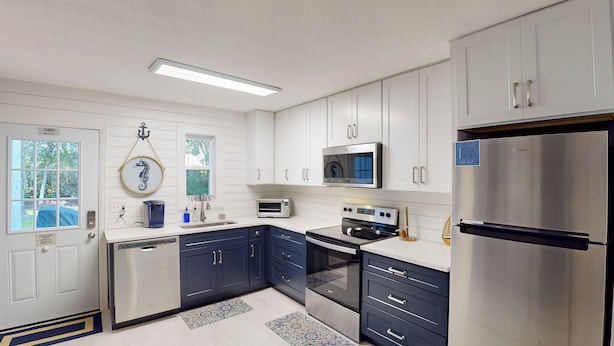 Una cocina moderna con armarios blancos y azul marino, electrodomésticos de acero inoxidable y un ventanal con vista a árboles.