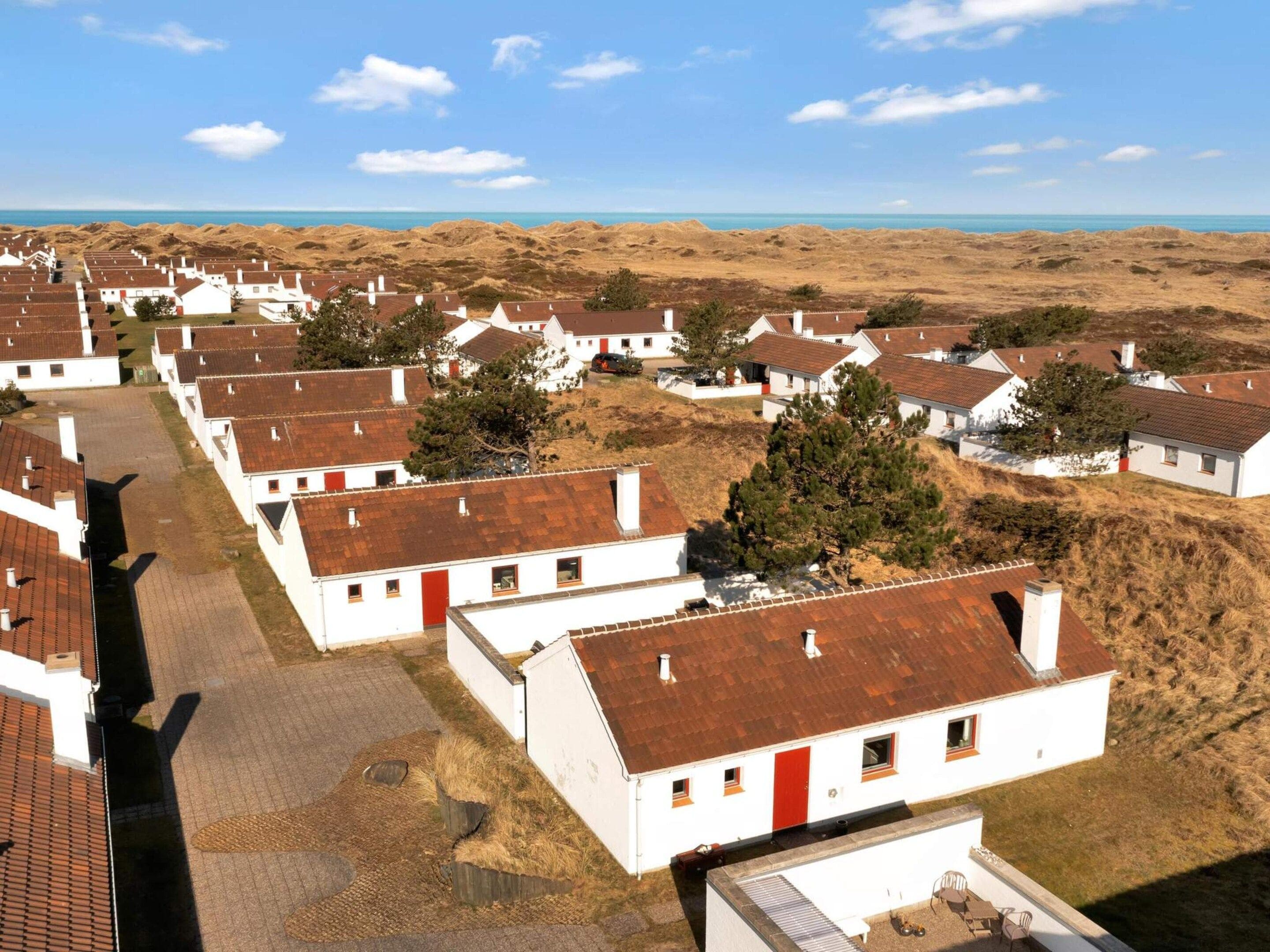 Un pueblo costero con casas blancas y puertas rojas, dispuesto en filas, con una playa de arena y el océano al fondo.