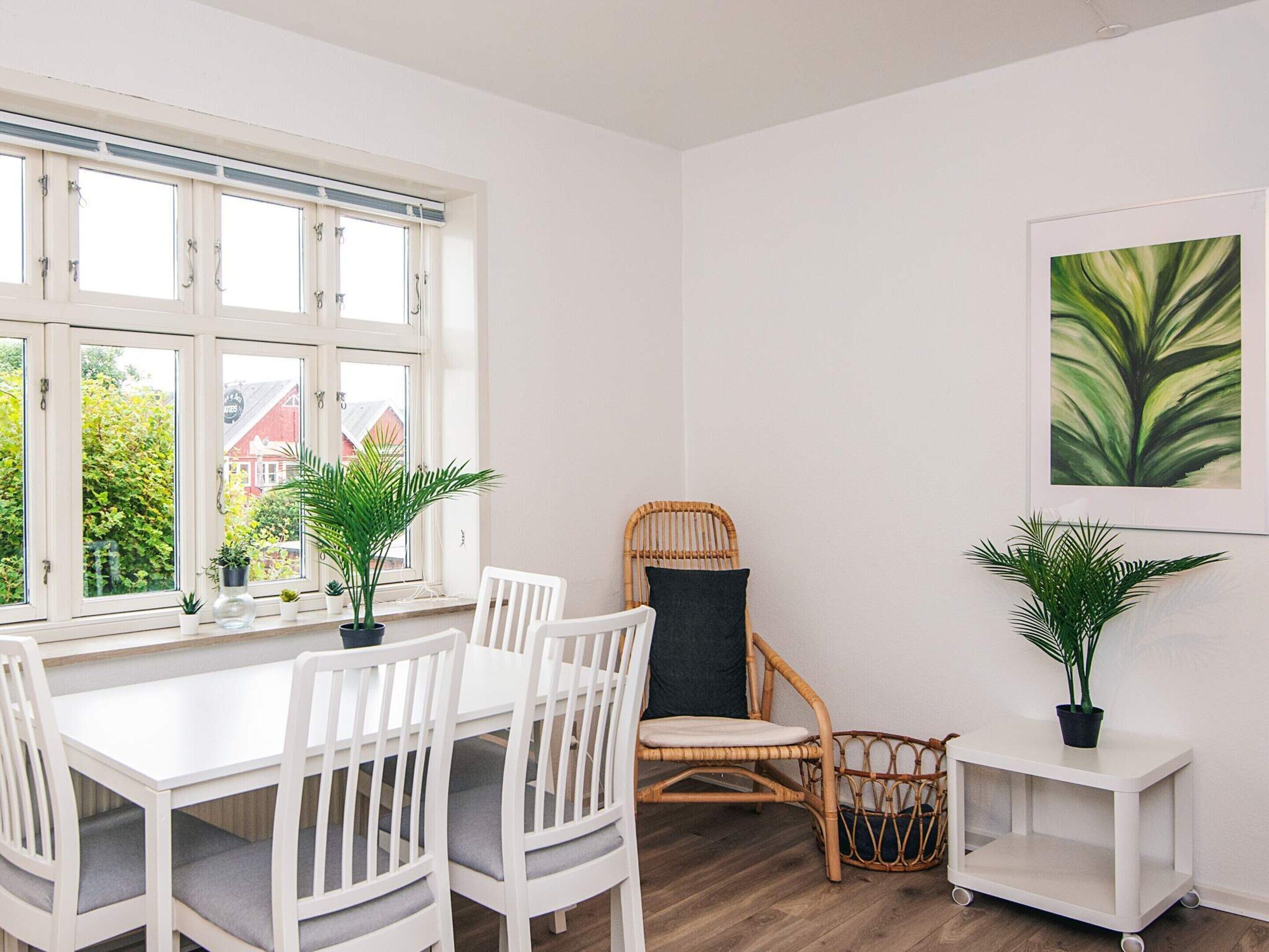 Une pièce lumineuse avec une table et des chaises blanches, un fauteuil en osier, une petite armoire blanche et une plante décorative accrochée au mur.
