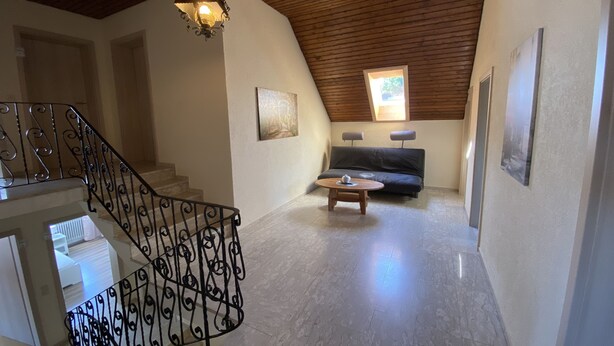 Un loft espacioso con techo de madera, una escalera con barandillas decorativas, una zona de estar acogedora con un sofá y una mesa de centro, y un ventanal que proporciona luz natural.