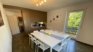 Un comedor moderno con una mesa y sillas blancas, una cocina al fondo y vista a vegetación a través de una puerta corredera.