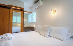 A hotel room with a bed, a wooden wardrobe, an air conditioning unit, a bedside lamp, and a window with blinds.