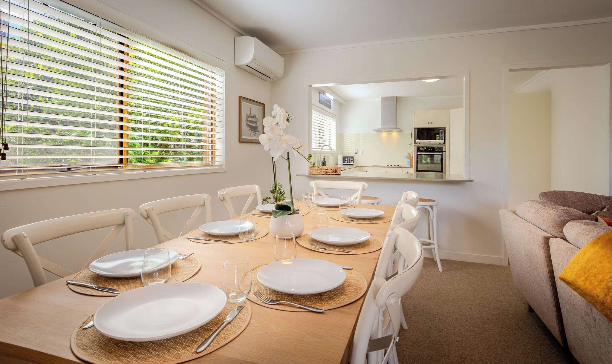 A dining area with a table set for six, white chairs, and a view of the kitchen through a window with blinds.