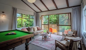A cozy room with a pool table, a sofa, and a chair, with large windows offering a view of greenery outside.