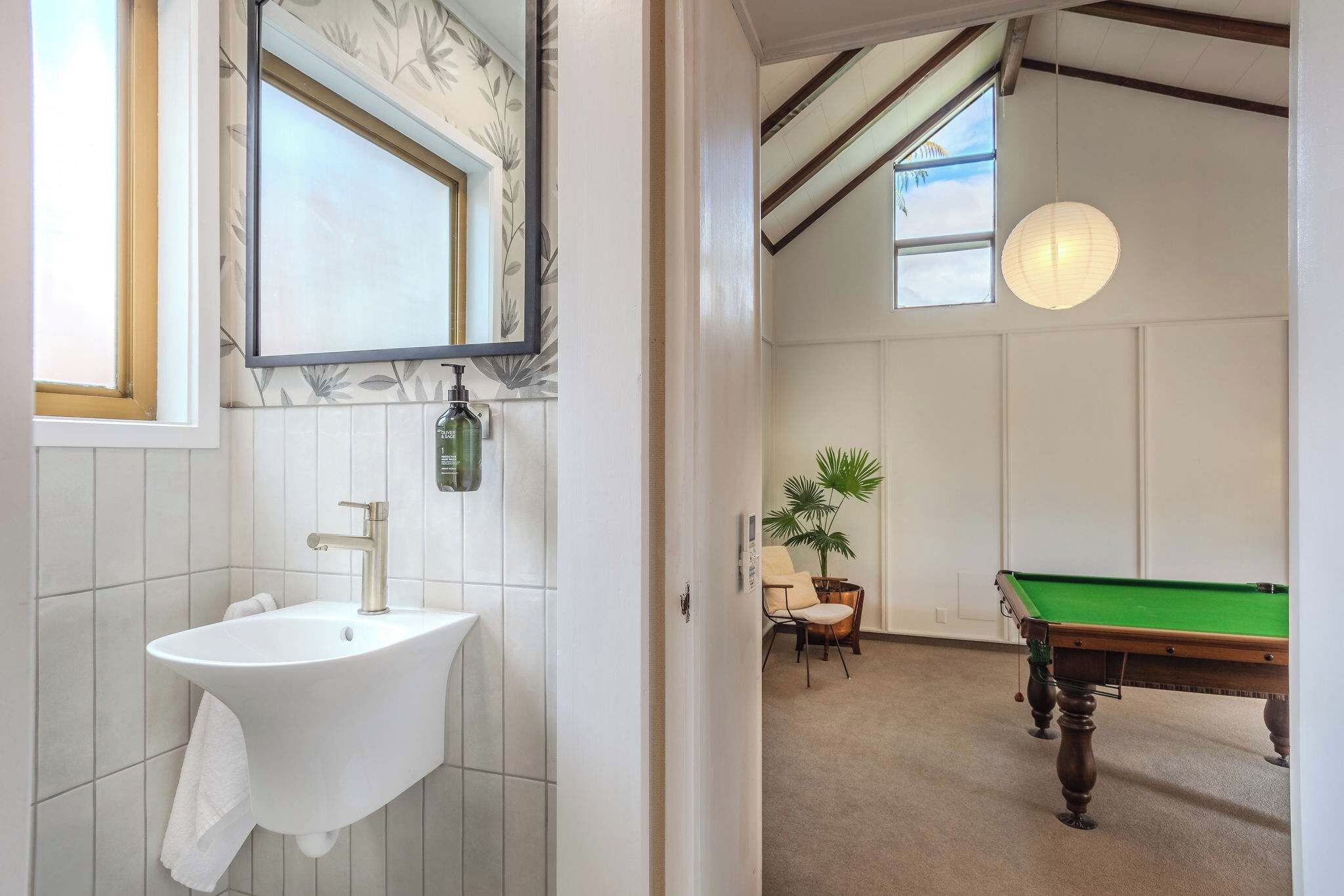 A bathroom with a sink, mirror, and a green pool table in the adjacent room.