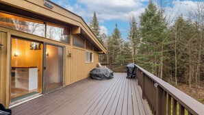 Eine Holzterrasse mit Glastür, einem überdachten Grillbereich und Blick auf einen bewaldeten Bereich.