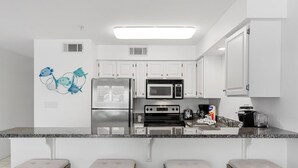Una cocina moderna con armarios blancos, un refrigerador de acero inoxidable, un microondas, un horno y una encimera de granito. Hay tres taburetes de bar con asientos acolchados.