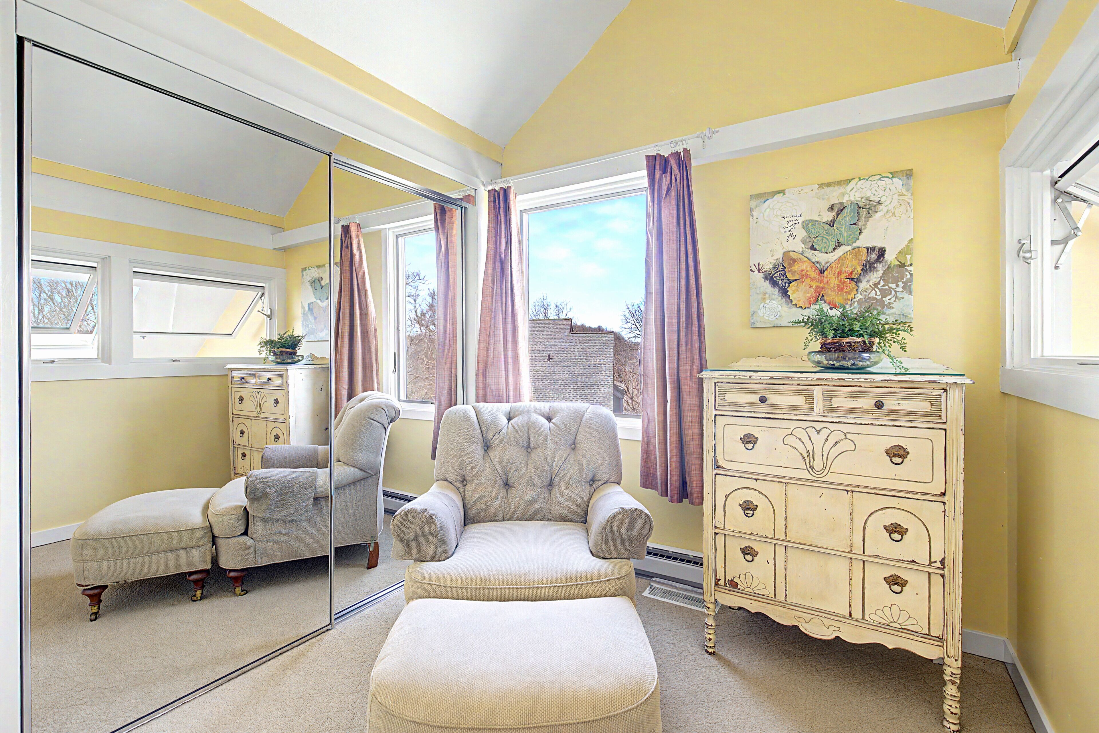 Quarto com parede espelhada, poltrona estofada, um banquinho combinando, cômoda e janela com cortinas.