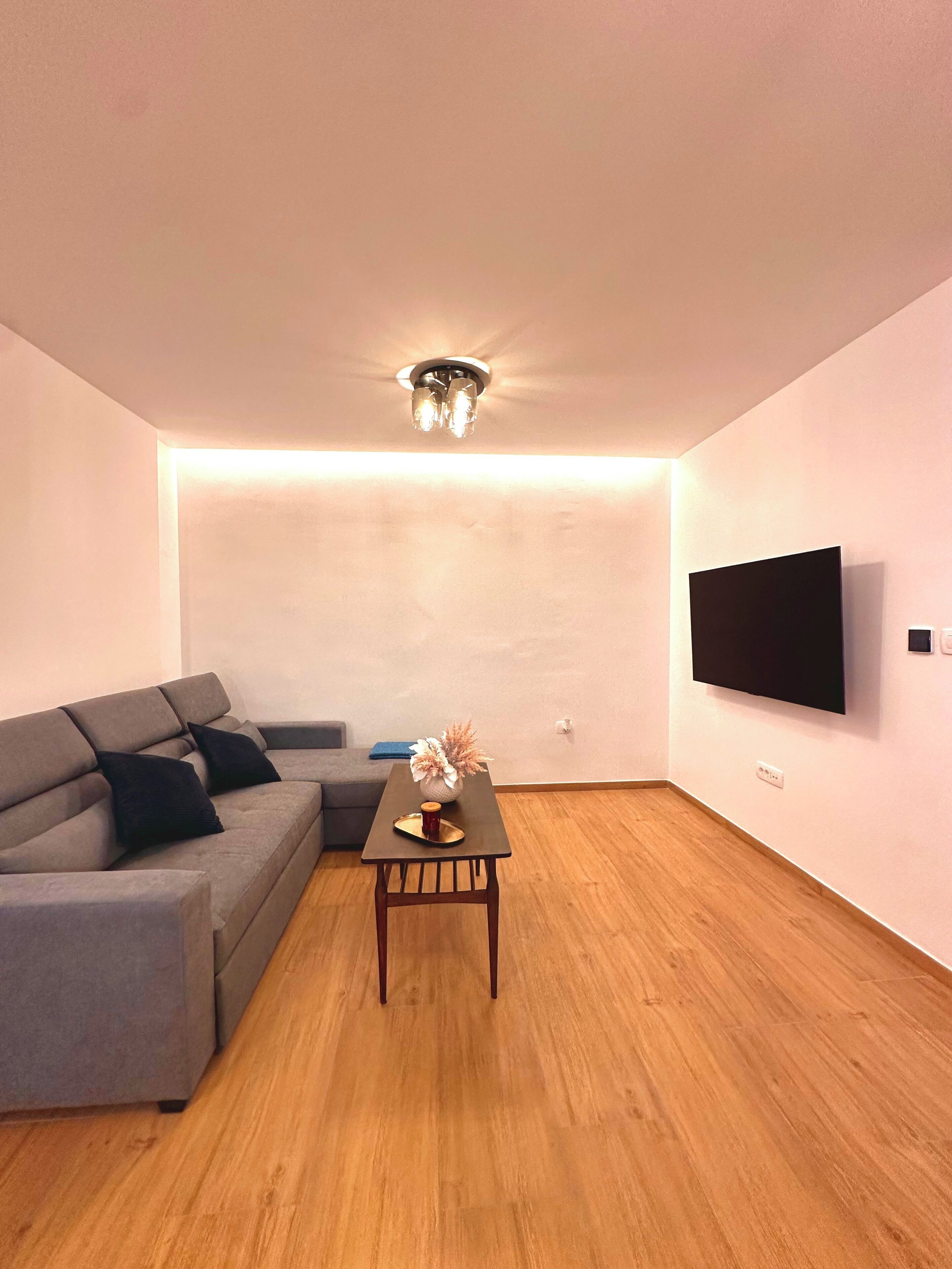 Un salon moderne avec un canapé d’angle gris, une table basse en bois et un téléviseur à écran plat fixé au mur.