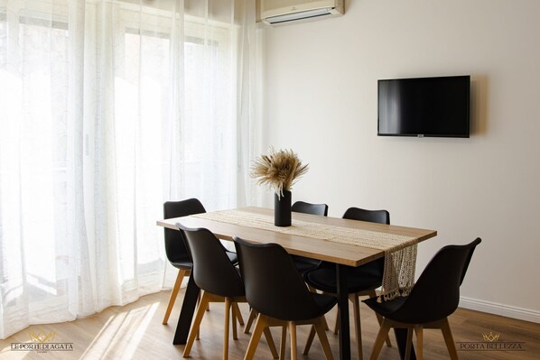 A dining area with a wooden table, black chairs, and a vase with dried plants. A flat-screen TV is mounted on the wall.