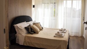 A neatly made bed with a black headboard, beige bedding, and several pillows. There are rolled towels on the bed, and the room has a wooden floor and a window with sheer curtains. - Holiday home catania 5km from the center relaxation apartment complete kitchen
 (Sicilia)