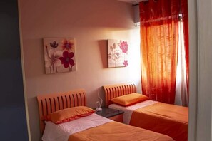 Habitación de hotel con dos camas, ropa de cama naranja, una mesita pequeña y dos cuadros colgados en la pared.