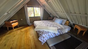 Um quarto de sótão aconchegante, com piso de madeira, uma cama com roupa de cama estampada, uma mesinha de cabeceira e uma janela com cortinas.