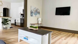 Una sala de estar moderna con piso de madera, una mesa de centro blanca con una cesta, un televisor de pantalla plana montado en la pared y una cocina visible al fondo.