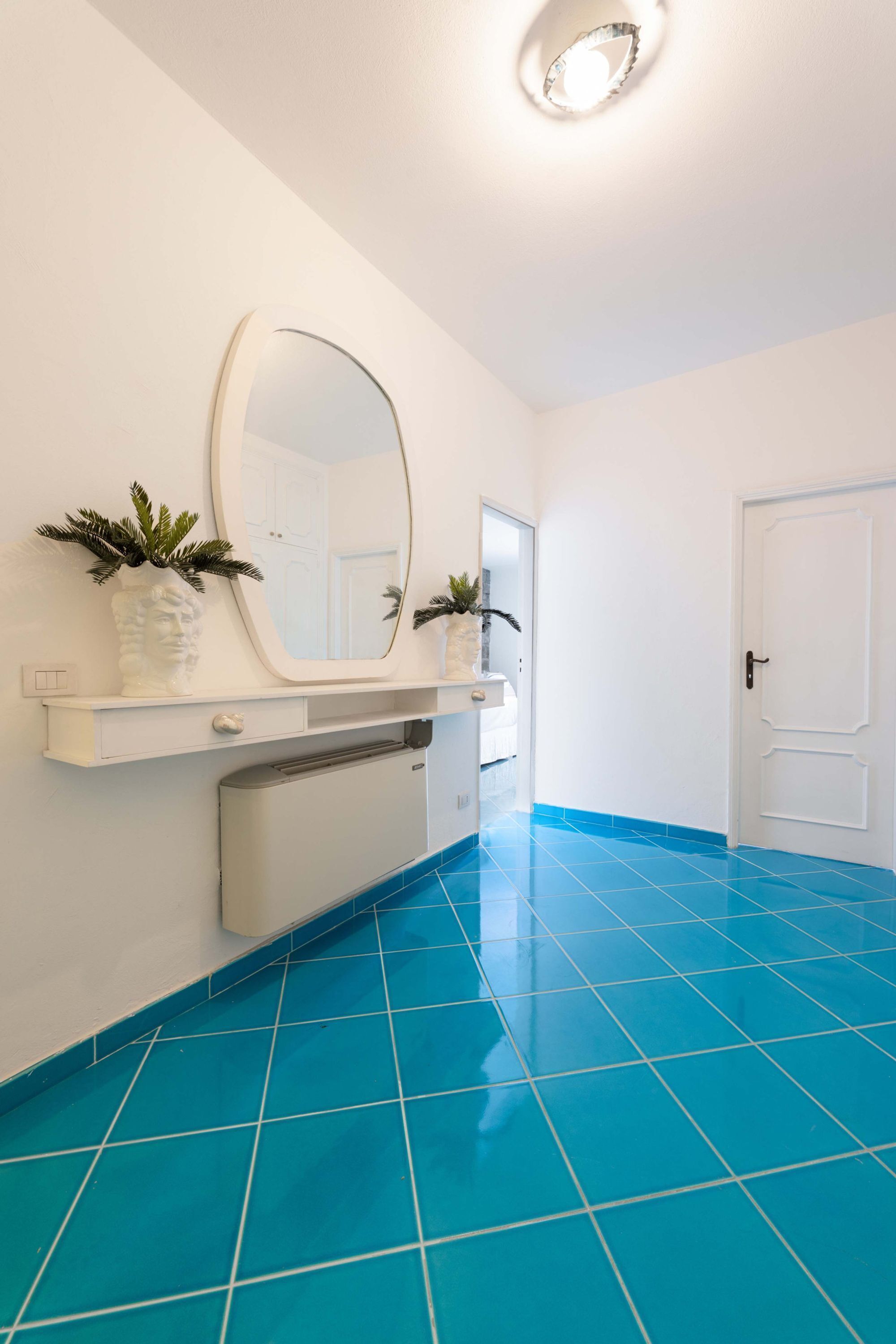 Un pasillo con una puerta blanca, un espejo blanco fijado a la pared y un estante blanco decorado con objetos.