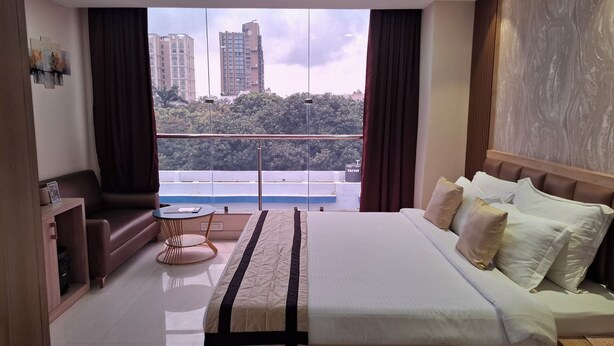 A hotel room with a large bed, a small round table, a sofa, and a view of tall buildings and trees through the window. - PARK EASTERN HOTEL (Kolkata)