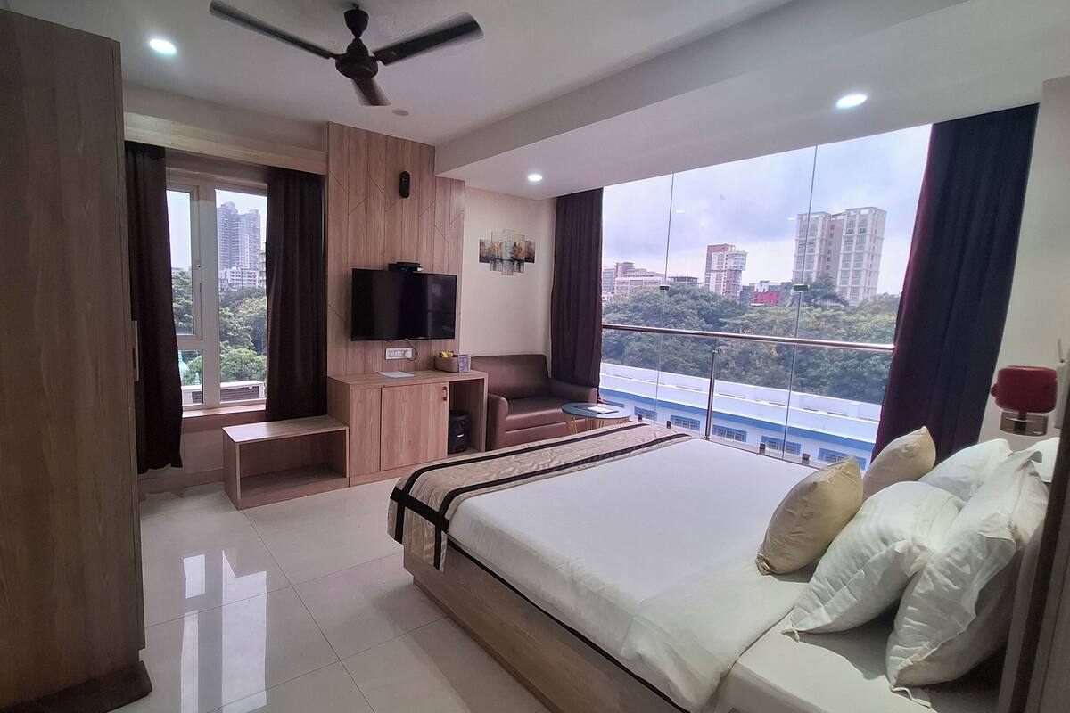 A modern hotel room with a large bed, a desk with a TV, a balcony with a view of the city, and a ceiling fan.