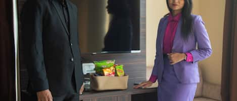 A man and a woman standing in a hotel room with a bed, a TV, and a small table with snacks.