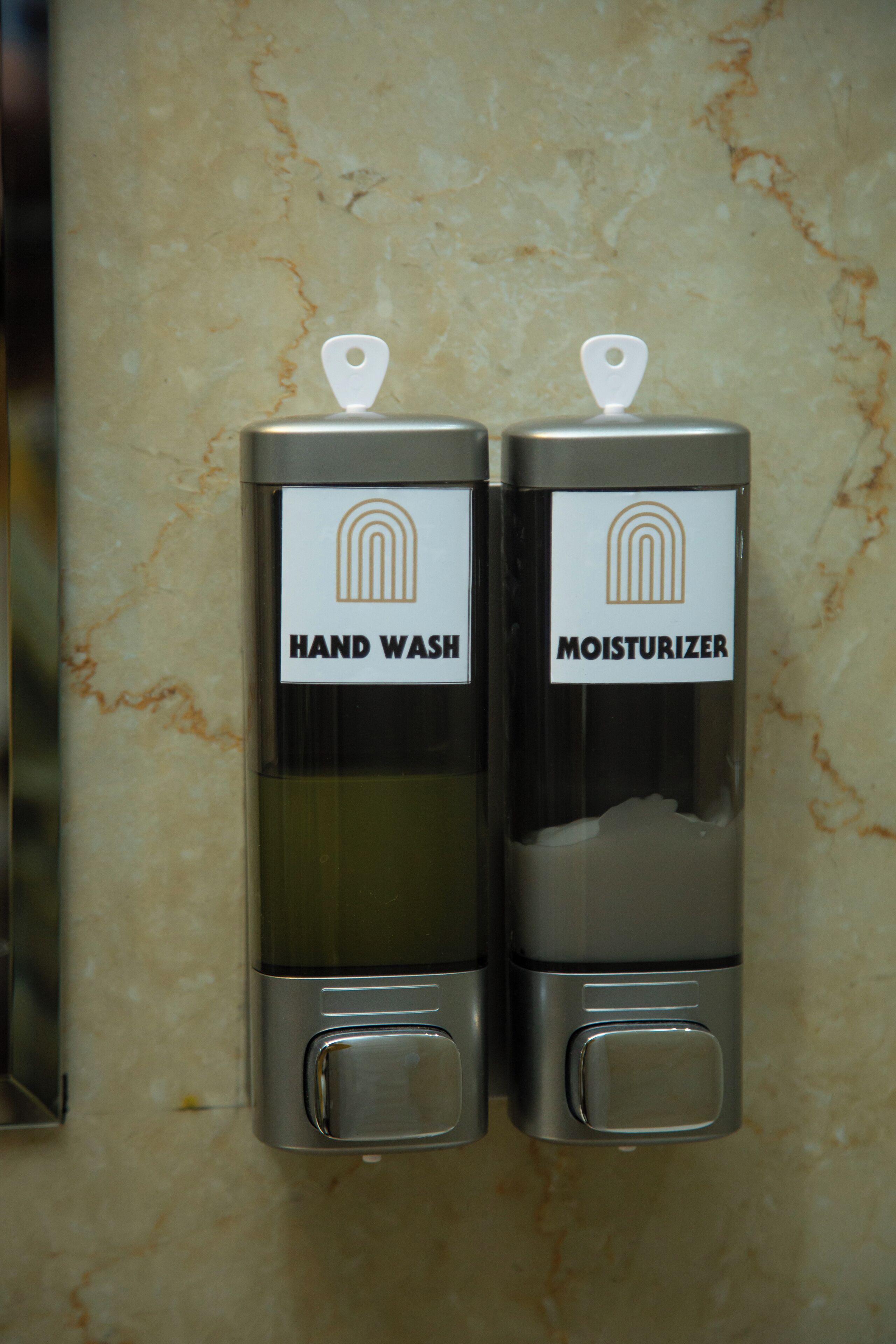 Zwei Spender mit den Aufschriften 'HAND WASH' und 'MOISTURIZER' an einer Marmorwand befestigt.