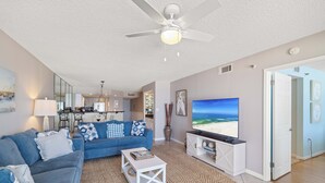 A living room with a blue sofa, a coffee table, a TV displaying a beach scene, a ceiling fan, and a kitchen in the background. - Summerchase 307 by Liquid Life (Orange Beach)