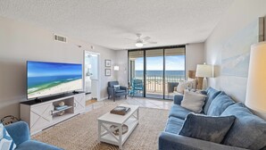 A living room with a television, a sofa, a coffee table, and a view of the beach through large glass doors. - Summerchase 307 by Liquid Life (Orange Beach)