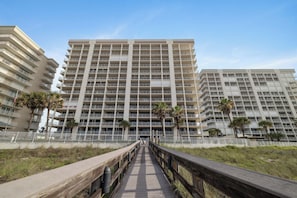 A multi-story hotel with a central walkway leading to the entrance, surrounded by palm trees and a white fence. - Windward Pointe 805 by Liquid Life (Orange Beach)