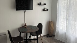 Una habitación de hotel moderna con televisor de pantalla plana, una mesa de comedor redonda con sillas, un estante decorativo con un cartel que dice 'LOVE', una lámpara de pie de mimbre y cortinas de gasa.