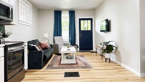 Ein modernes Wohnzimmer mit Sofa, Couchtisch und Fernseher. Eine Tür, ein Fenster mit Vorhängen und eine Küchenzeile mit Mikrowelle sind ebenfalls vorhanden.