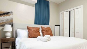 Ein ordentlich bezogenes Bett mit weißer Bettwäsche und zwei braunen Kissen, ein Nachttisch mit Lampe und ein blauer Vorhang über dem Bett.