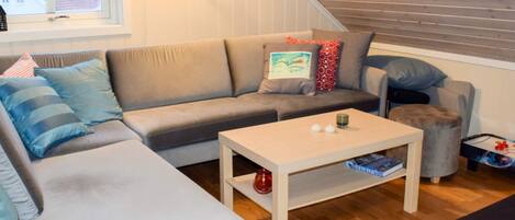 Ein gemütliches Wohnzimmer mit einem grauen Sofa, einem weißen Couchtisch und einem kleinen Hocker. Ein Fenster mit Herz-Dekoration ist sichtbar, und es gibt eine Holzzieldecke.