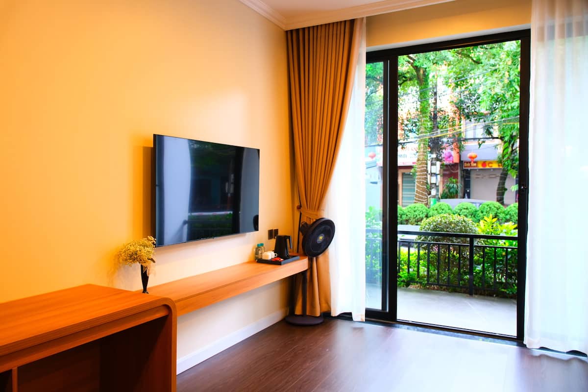 A room with a flat-screen TV mounted on the wall, a wooden console table with a vase of flowers, a standing fan, and a sliding glass door leading to a balcony with greenery outside.