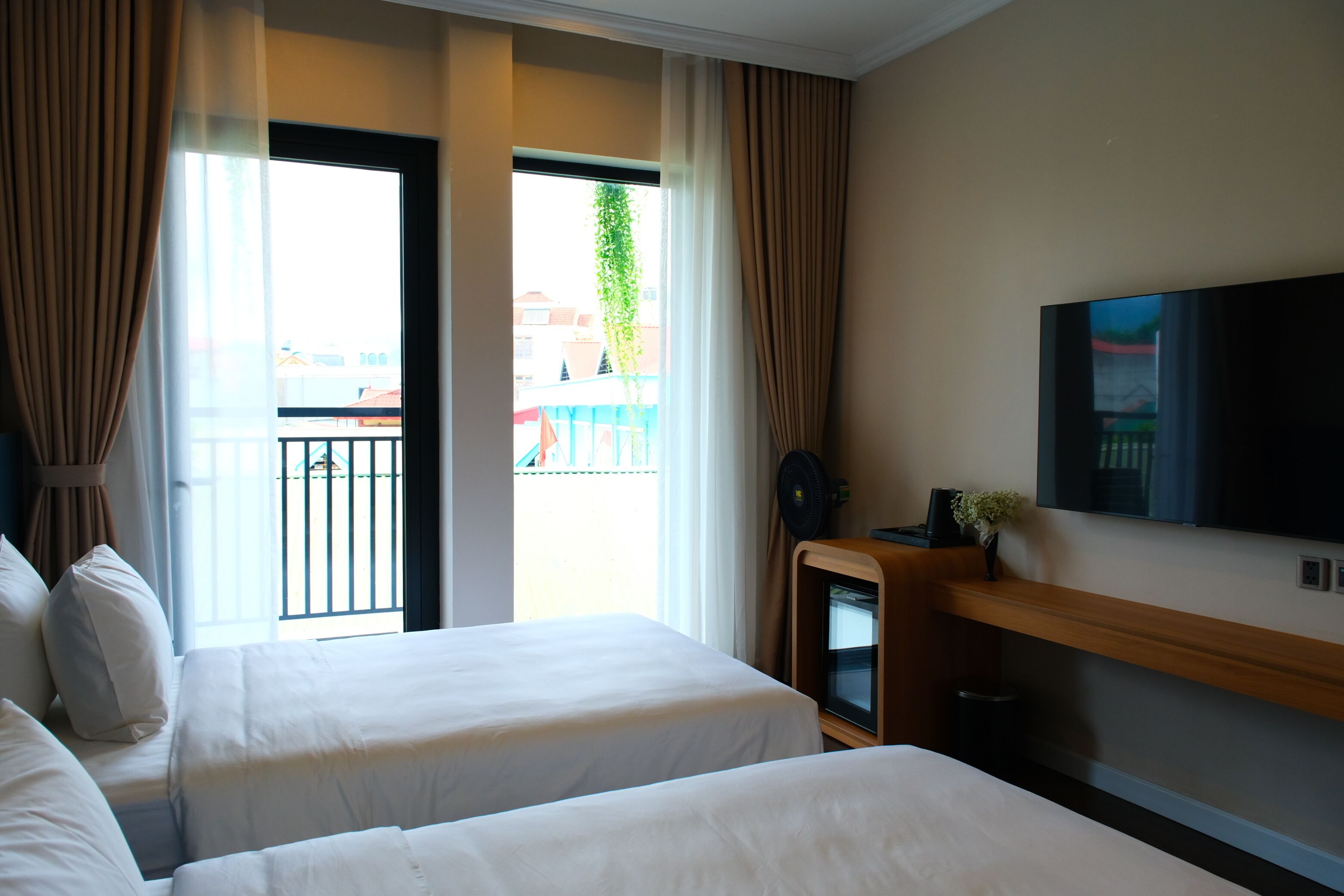 A hotel room with two beds, a TV, a small table with a plant, and a balcony with a view of buildings and greenery.