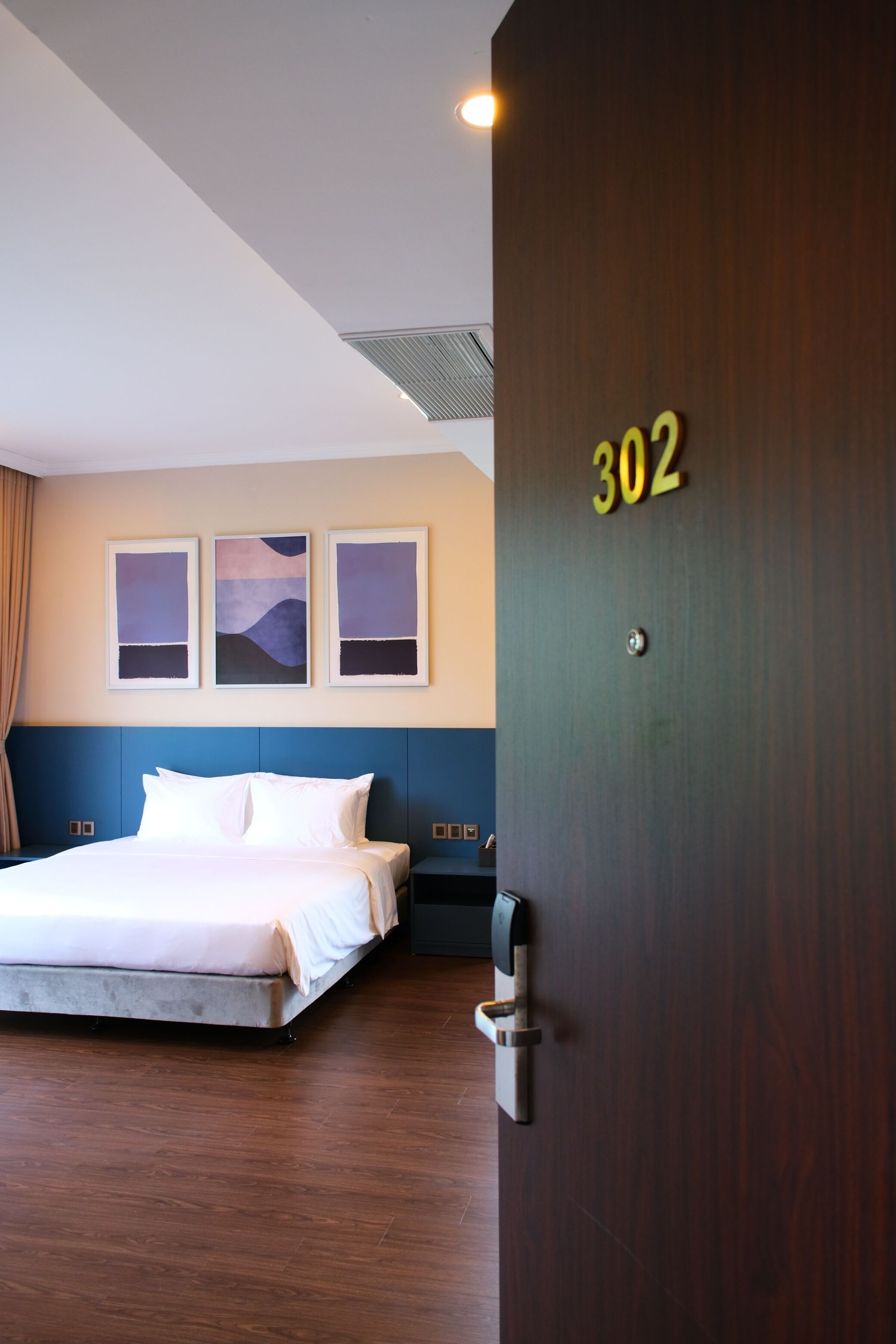 Quarto de hotel com uma cama, uma porta com o número 302 e três quadros na parede.