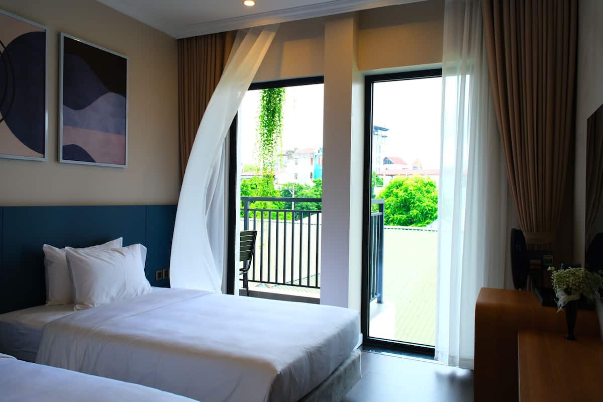 A hotel room with a bed, a desk, and a balcony with a view of greenery and buildings.