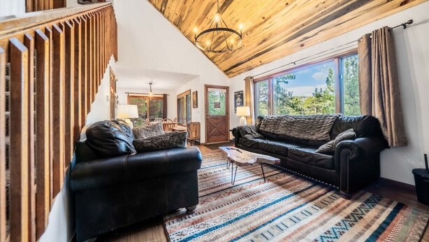 A spacious living room with a high ceiling, wooden beams, a black leather sofa, a coffee table, and a large window offering a view of trees. - Walk Into National Park! River Access & Fishing (Estes Park)