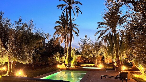 Un área de piscina al aire libre de noche, con una piscina rectangular, sillas de descanso y palmeras.