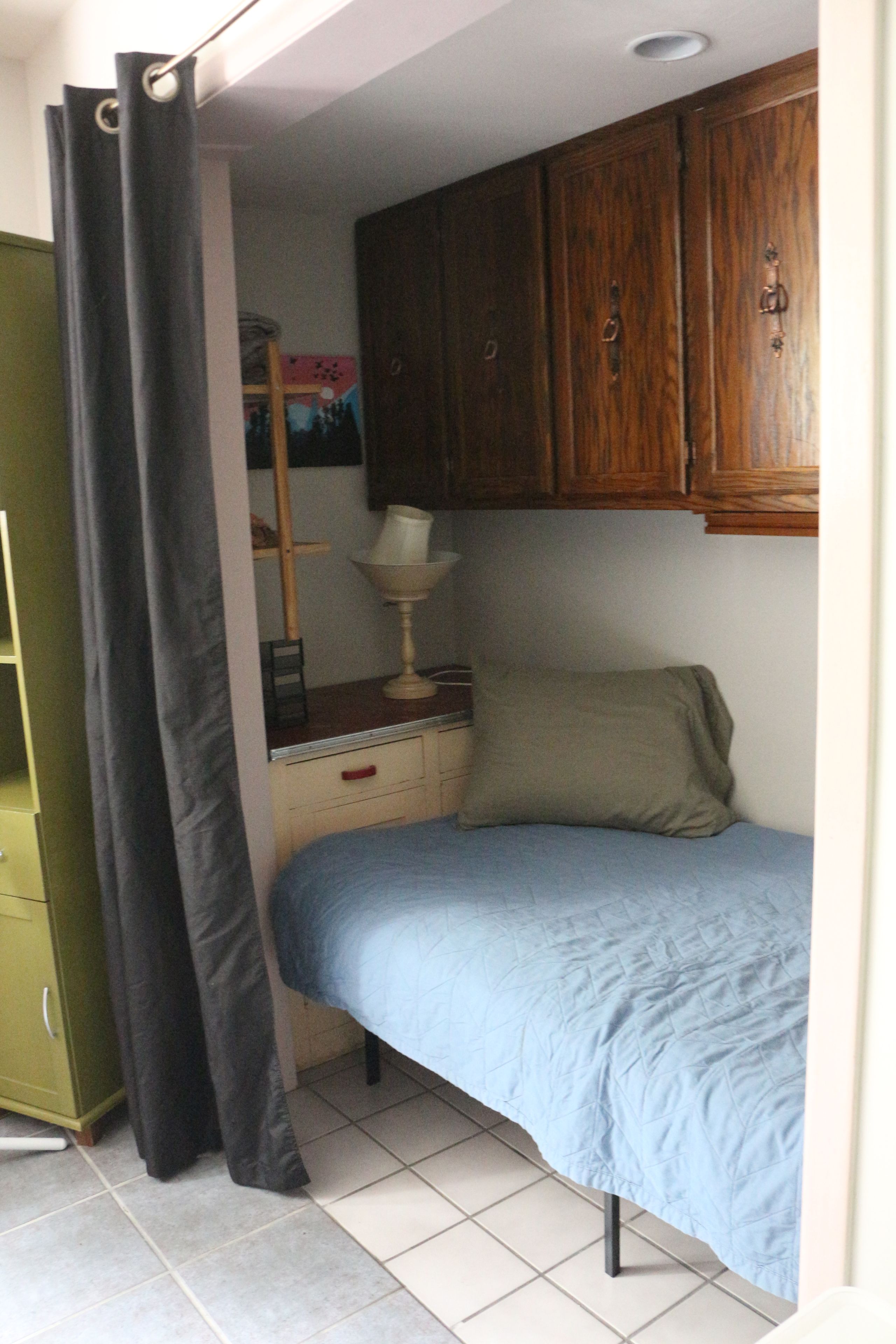 A small, single-bed room with a wooden cabinet above the bed, a bedside table with a lamp, and a green wardrobe.