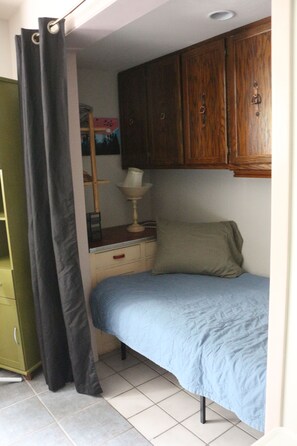 A small, single-bed room with a wooden cabinet above the bed, a bedside table with a lamp, and a green wardrobe.