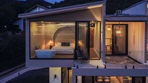 Une maison moderne à deux étages dotée d’une chambre à coucher entièrement vitrée, comprenant une grande baignoire, un espace de vie séparé avec un canapé et des fauteuils, ainsi qu’un balcon.