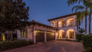 A two-story house with a driveway leading to a garage, surrounded by palm trees and a well-maintained lawn.