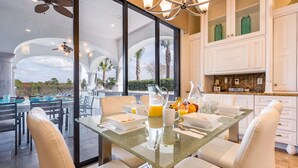 A modern dining area with a glass table, white chairs, and a view of palm trees through large windows.