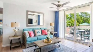 Ein Wohnzimmer mit einem blauen Sofa, einem Couchtisch und einem Deckenventilator. Eine Glastür öffnet sich zu einem Außenbereich mit Tisch und Stühlen.