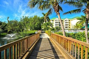A wooden boardwalk leading to a hotel with a green roof, surrounded by lush greenery and palm trees. - The Butterfly Nest | Newly Updated w/ Shared Pool (Key West)